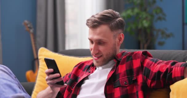Närbild av unga glada leende man skriva meddelande via smartphone medan du sitter på modern interiör — Stockvideo