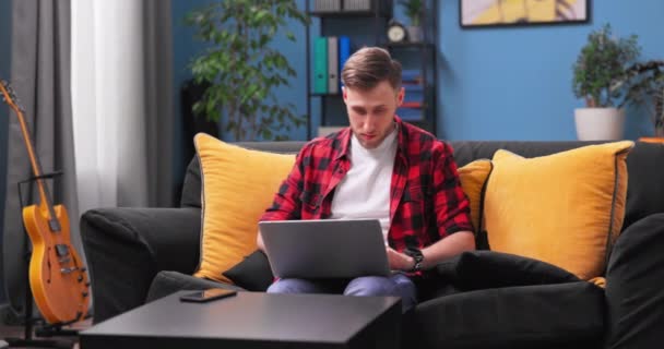 Усміхнений хлопчик тисячоліття сидить на дивані, використовуючи сучасний ноутбук, переглядаючи необмежений бездротовий Інтернет, щасливий — стокове відео