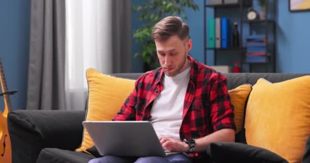 Усміхнений молодий чоловік використовує ноутбук, сидячи на дивані вдома, красиві чоловічі покупки або балачки — стокове відео