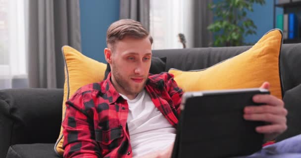 Усміхнений молодий студент, використовуючи цифровий планшет вдома, тисячолітній користувач тримає комп'ютер — стокове відео