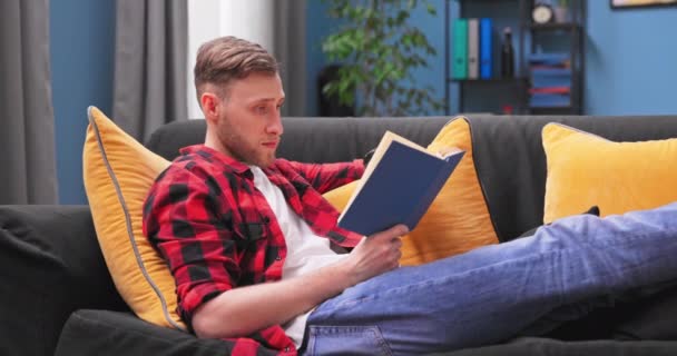 Šťastný tisíciletý chlapec ležel na gauči doma a četl zajímavou knihu, klidný americký úsměv — Stock video