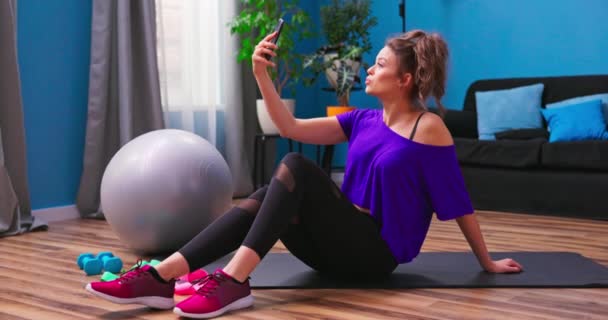 En söt ung kvinna klädd i fitness kläder sitter på trägolvet i en — Stockvideo