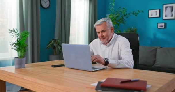 Serious senior man sit at cozy home office table working on laptop, 55s businessman consider — Vídeo de stock