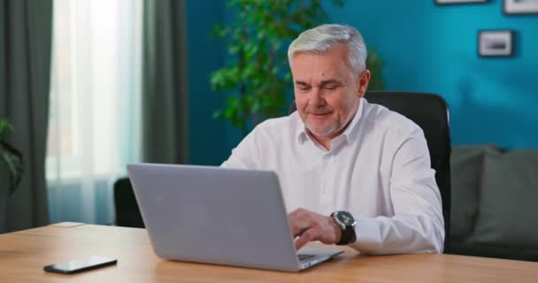 Серйозний старший чоловік сидить за затишним домашнім офісним столом, працюючи на ноутбуці, 55-річний бізнесмен вважає — стокове відео