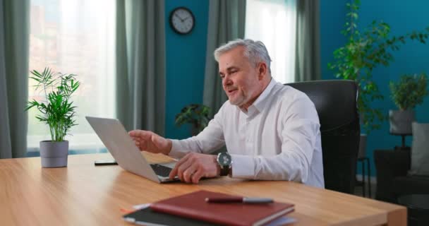 Щасливий зрілий 55-річний чоловік сидить за столом вдома, дивлячись на екран пристрою, використовуючи відеозапис — стокове відео