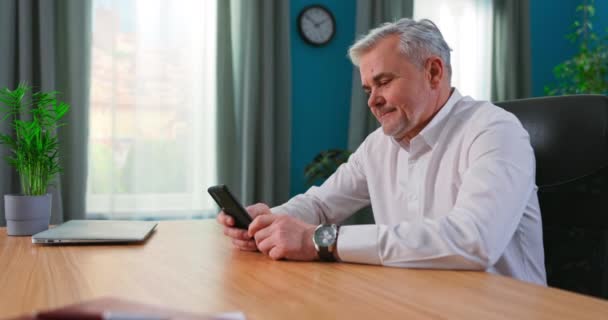 Мужчина средних лет 55 сидит в помещении, держа мобильный телефон смс улыбки наслаждаться легко онлайн — стоковое видео