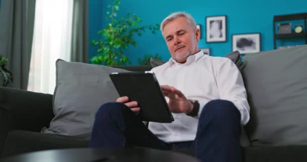 Hombre maduro sonriente utilizando tableta digital tableta moderna tecnología relajarse en el sofá. Feliz jubilado — Vídeo de stock
