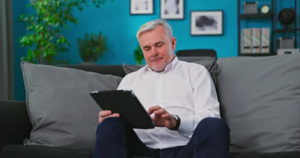 Hombre caucásico mayor sonriendo, viejo hombre de mediana edad sentado en el sofá usando tableta digital en — Vídeo de stock