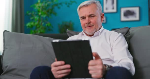 Sonriente anciano de mediana edad relajante celebración tableta digital lectura e libro sentarse en el sofá — Vídeos de Stock