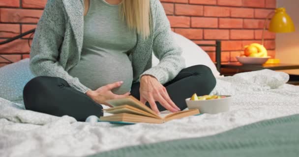Крупный план беременной девушки, читающей книгу, ласкающей живот, сидящей — стоковое видео