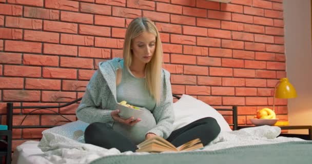 Mooie zwangere vrouw leest boek zittend in bed thuis en glimlachend genieten van literatuur. — Stockvideo