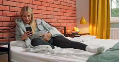 Genç hamile bir kadın, kocasına akıllı telefondan mesaj atarak yatakta dinleniyor. Güzel, zarif bayan.