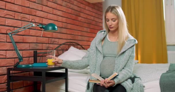 Dobře vypadající těhotná žena sedí na útulné posteli čtení knihy, snění, pití šťávy — Stock video