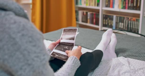 Видео красивой молодой беременной женщины, смотрящей ультразвуковые отпечатки во время лежания на кровати. — стоковое видео