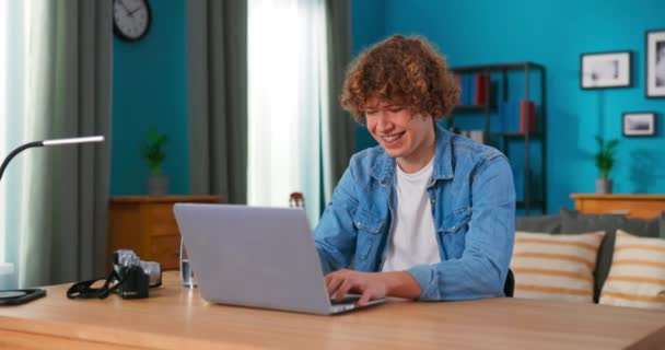 Guapo adolescente con el pelo rizado sentado en un escritorio en una sala de estar y — Vídeos de Stock
