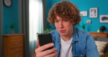 Gülümseyen çekici genç çocuk evde masada oturmuş akıllı telefon kullanarak arkadaşlarıyla sohbet ediyor.