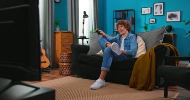 Kanallarda sörf yapmak. Neşeli genç Avrupalı beyaz erkek televizyon izliyor ve uzaktan kumandayı elinde tutuyor