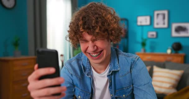Миллениальный красавчик с помощью смартфона смотрит в камеру и делает смешные гримасы веселящимися — стоковое видео