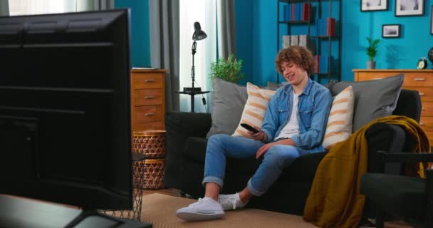 Вид бородатого подростка, сидящего на диване и смотрящего телевизор — стоковое видео