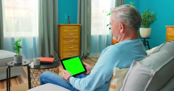 Homem mais velho em casa sentado em um sofá funciona em um tablet com Green Mock-up — Vídeo de Stock