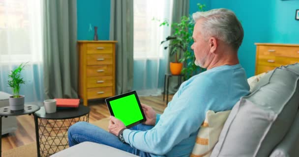 Adulto anciano en casa utiliza Green Mock-up Screen Tablet. Él está sentado en un — Vídeo de stock
