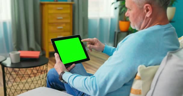 Adulto anciano en casa utiliza Green Mock-up Screen Tablet. Él está sentado en un — Vídeo de stock