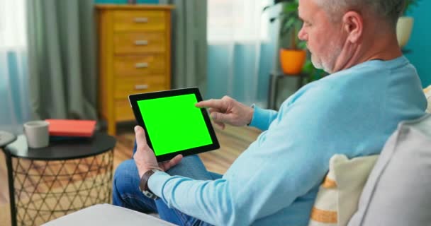 Starszy mężczyzna w domu używa zielonego ekranu. Siedzi na kanapie. — Wideo stockowe