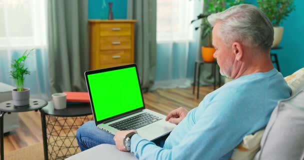 Senior Man Λειτουργεί σε έναν φορητό υπολογιστή με πράσινη οθόνη Mock-up κάθεται σε έναν καναπέ — Αρχείο Βίντεο