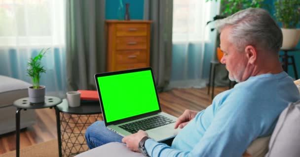 Homem mais velho em casa sentado em um sofá funciona em um computador portátil com verde — Vídeo de Stock