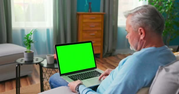 Homem mais velho em casa sentado em um sofá funciona em um computador portátil com verde — Vídeo de Stock
