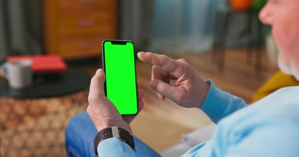 Hombre mayor en casa utiliza el teléfono celular de pantalla falsa verde. Él está sentado en un — Vídeo de stock