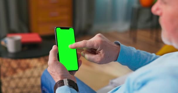 Взрослый старик дома использует зеленый макет экрана смартфона. Он сидит на — стоковое видео
