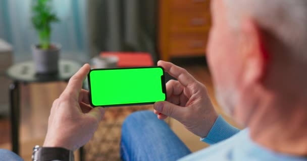 Älterer Mann zu Hause auf der Couch mit grünem Bildschirm-Smartphone — Stockvideo