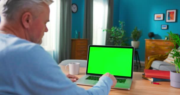 Hombre mayor trabaja en una computadora portátil con Green Mock-up Screen sentado en el escritorio en — Vídeos de Stock