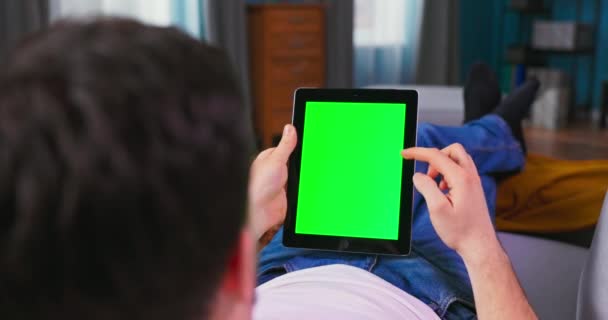 Jovem em casa descansando em um sofá usando com o computador tablet tela Mock-up verde — Vídeo de Stock