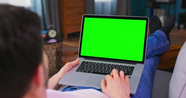 Homem em casa deitado em um sofá trabalhando no computador portátil com tela de Mock-up verde. — Vídeo de Stock