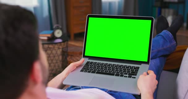 Νεαρός στο σπίτι χρησιμοποιεί πράσινο mock-up οθόνη laptop. Είναι ξαπλωμένος στον καναπέ. — Αρχείο Βίντεο