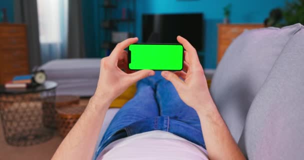 Joven en casa acostado en un sofá usando con Green Mock-up Screen Smartphone. Muchacho — Vídeo de stock
