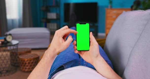 Homem em casa deitado em um sofá usando Smartphone com tela de Mock-up verde, fazendo Swiping, — Vídeo de Stock