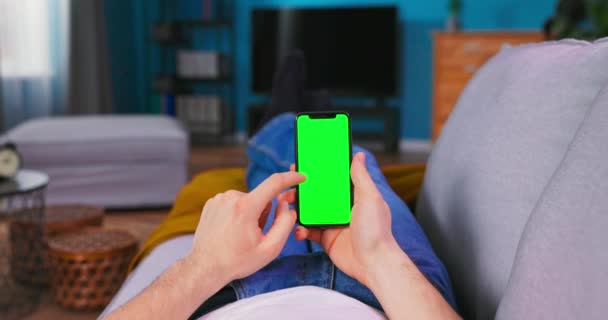 Человек дома лежит на диване, используя смартфон с зеленым макетом экрана, делает Swiping, — стоковое видео