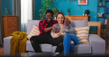 Neşeli Kafkasyalı genç çift, birlikte kanepede televizyon izlerken gülüp sohbet ediyorlar.