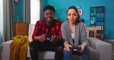 Neşeli melez çiftler, Afrika kökenli Amerikalı erkekler ve beyaz kadınlar kanepede oturup joysticklerle video oyunları oynuyorlar. Neşeli kadın, oyunda erkeği yenerken eğleniyor..
