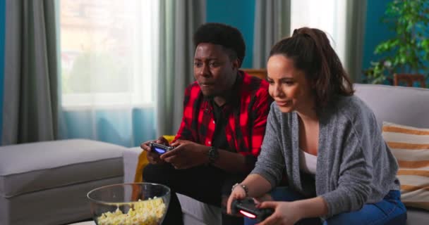 Mladý šťastný mezinárodní pár sedí doma na gauči a hraje videohry na konzoli. — Stock video