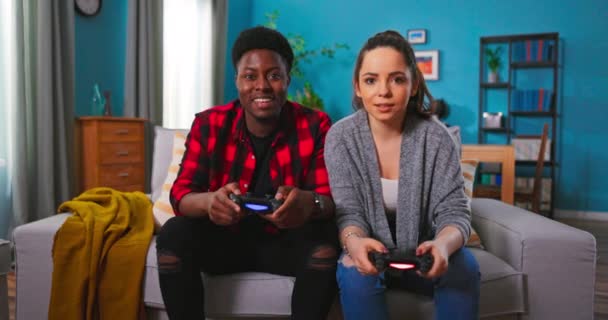 Glada blandraser par afroamerikanska Man och kaukasisk kvinna vilar i rummet sitter på soffan spelar videospel med hjälp av joysticks. Glada kvinna som har kul vinna över mannen i spelet. — Stockvideo