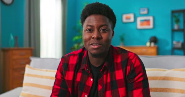 En smuk afrikansk amerikansk mandlig studerende sidder på sofaen i en stue og er vært – Stock-video