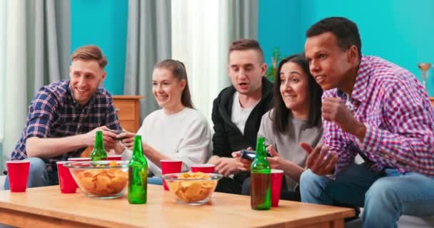 多民族の人々は自宅のリビングルームでビデオゲームの勝利を祝います。ビールやビールを楽しむ多様な友人たち — ストック動画