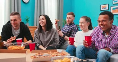 Neşeli arkadaşlar evde pizza yiyorlar. Mutlu gülümseyen erkekler ve genç kadınlar pizzayı paylaşıyor