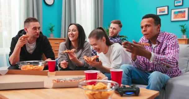 Los jóvenes alegres hombres y mujeres están comiendo pizza cursi, charlando relajante durante la fiesta de interior — Vídeos de Stock