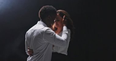 Film sahnesi, tiyatro oyunu, aşk oyunculuğu, iki insanın şefkatli selamlaşması, sevgilisini kucaklayan bir adam, saçını tarayan bir kız, parmaklarını partnerinin dudaklarında gezdiriyor, buruna dokunuyor, gülümsüyor, dinç, siyah bir arkaplan.