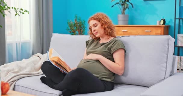 La futura madre se sienta en el sofá de la sala de estar y se relaja leyendo un libro para bebés, — Vídeos de Stock